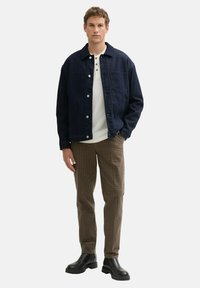 Dunkle Jeansjacke mit Knöpfen, weißes Hemd darunter, braune karierte Hose und schwarze Stiefeletten. Einfaches Design mit legerer Passform.