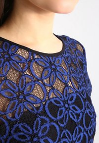 Žena v modrém květinovém krajkovém topu přes černou látku, odhalující dekolt a ramena s pokožkou viditelnou skrz krajkový vzor.