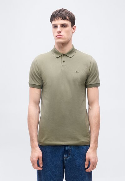 Jeune homme portant un polo à manches courtes vert olive avec un petit logo "BOSS" sur la poitrine et un jean bleu, debout devant un fond uni.