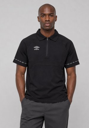 Mann trägt ein schwarzes Polo-Shirt mit kurzen Ärmeln und Umbro-Logo sowie graue Hose, steht mit Händen in den Taschen vor einfachem Hintergrund.