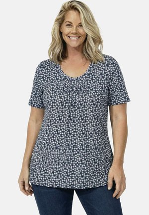 Lächende Frau mit schulterlangem blondem Haar, die ein marineblaues kurzärmeliges Oberteil mit kleinem weißem Blumenmuster und dunkelblaue Jeans trägt.
