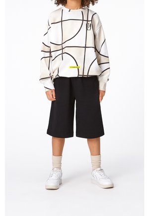 Personne portant un pull beige à motif abstrait, un short noir mi-genoux, des chaussettes beiges et des baskets blanches, debout devant un fond blanc.