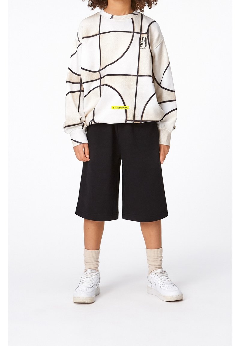 Personne portant un pull beige à motif abstrait, un short noir mi-genoux, des chaussettes beiges et des baskets blanches, debout devant un fond blanc.