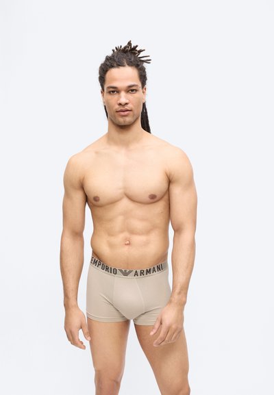 Μυώδης άνδρας με dreadlocks φοράει μπεζ σλιπ boxer Emporio Armani, στέκεται μπροστά σε απλό ανοιχτό φόντο με ουδέτερη έκφραση.