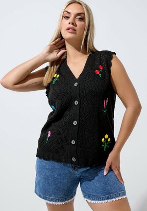 Donna con lunghi capelli biondi indossa un gilet nero senza maniche lavorato a maglia con ricamo floreale colorato e pantaloncini di denim blu con bordura in pizzo bianco.