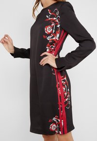 Zwarte jurk met lange mouwen, een ronde halslijn, rode bloemenpatronen aan de zijkant en rode accentstroken. Rechte stofstructuur.
