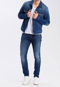 Denim jacket in dark blue with buttoned chest pockets, worn over a white V-neck shirt, paired with slim-fit blue jeans and white sneakers.