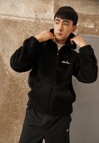 Svart fleece zip-up jakke med hette, med en logo på brystet og en teksturert overflate. Kombinert med svarte bukser.