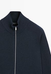 Pull en tricot bleu marine foncé avec fermeture éclair, col côtelé haut et zip argenté, montrant le haut du torse et les détails des épaules.