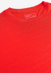 T-shirt rouge à manches courtes en tissu léger, avec un col rond et une texture subtile. L'étiquette indique "NEXT ACTIVE, FABRIQUÉ AU CAMBODGE."