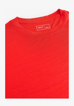 Rode t-shirt met korte mouwen gemaakt van lichtgewicht stof, met een ronde halslijn en subtiele textuur. Etiket leest "NEXT ACTIVE, MADE IN CAMBODIA."