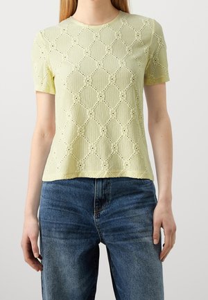 Top de punto texturizado en amarillo pálido de manga corta con patrón de ojales de diamantes y flores, combinado con vaqueros azules.
