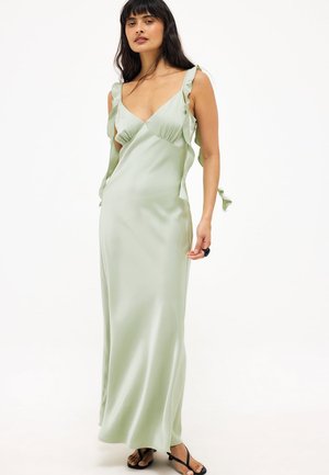 Kobieta w bladozielonej satynowej sukience na ramiączkach z falbankami, stojąca boso na jednolitym białym tle.