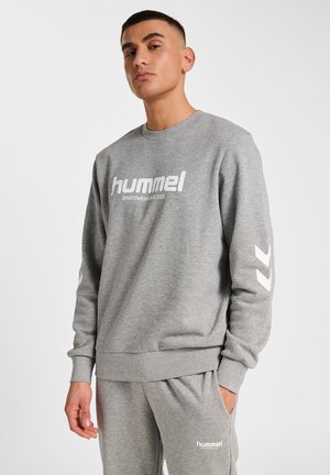 Man in grijze hummel sportkleding sweatshirt en bijpassende broek met logo tekst en chevron details op de mouwen, staande tegen een witte achtergrond.