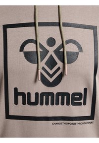 Beige hoodie med en svart grafisk logotyp. Har dragsko med beige spetsar. Texten under logotypen lyder "FÖRÄNDRA VÄRLDEN GENOM SPORT."