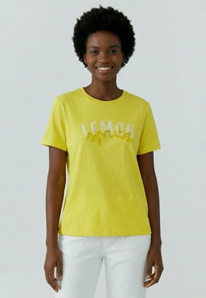Lächelnde Person mit lockigem Haar, die ein gelbes T-Shirt mit „LEMON“-Aufschrift und weiße Hosen trägt, steht vor einem einfarbigen hellen Hintergrund.