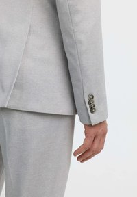 Hellegrauer Sakkoschirm mit fünf dunklen Knöpfen. Das Material wirkt glatt und strukturiert, mit subtilen Schneiderdetails und klaren Linien.