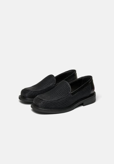 Ζευγάρι μαύρα πλεκτά δερμάτινα loafers χωρίς κορδόνια με στρογγυλεμένες μύτες και χαμηλά τακούνια σε απλό ανοιχτό φόντο.