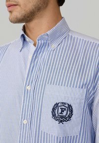 Camicia button-up a righe light blue e bianche con taschino sul petto che presenta un emblema ricamato e l'anno "1950." Tessuto in cotone, design con colletto.