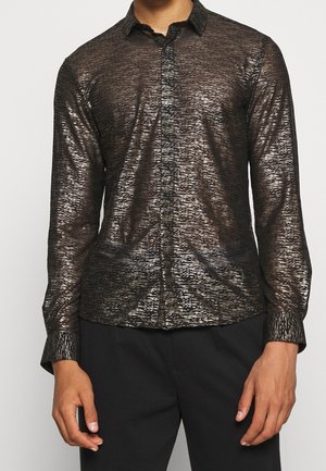 Mężczyzna ubrany w dopasowaną, zapinaną na guziki koszulę z długim rękawem o wzorze czarnych i metalicznych poziomych pasków, w połączeniu z czarnymi spodniami.