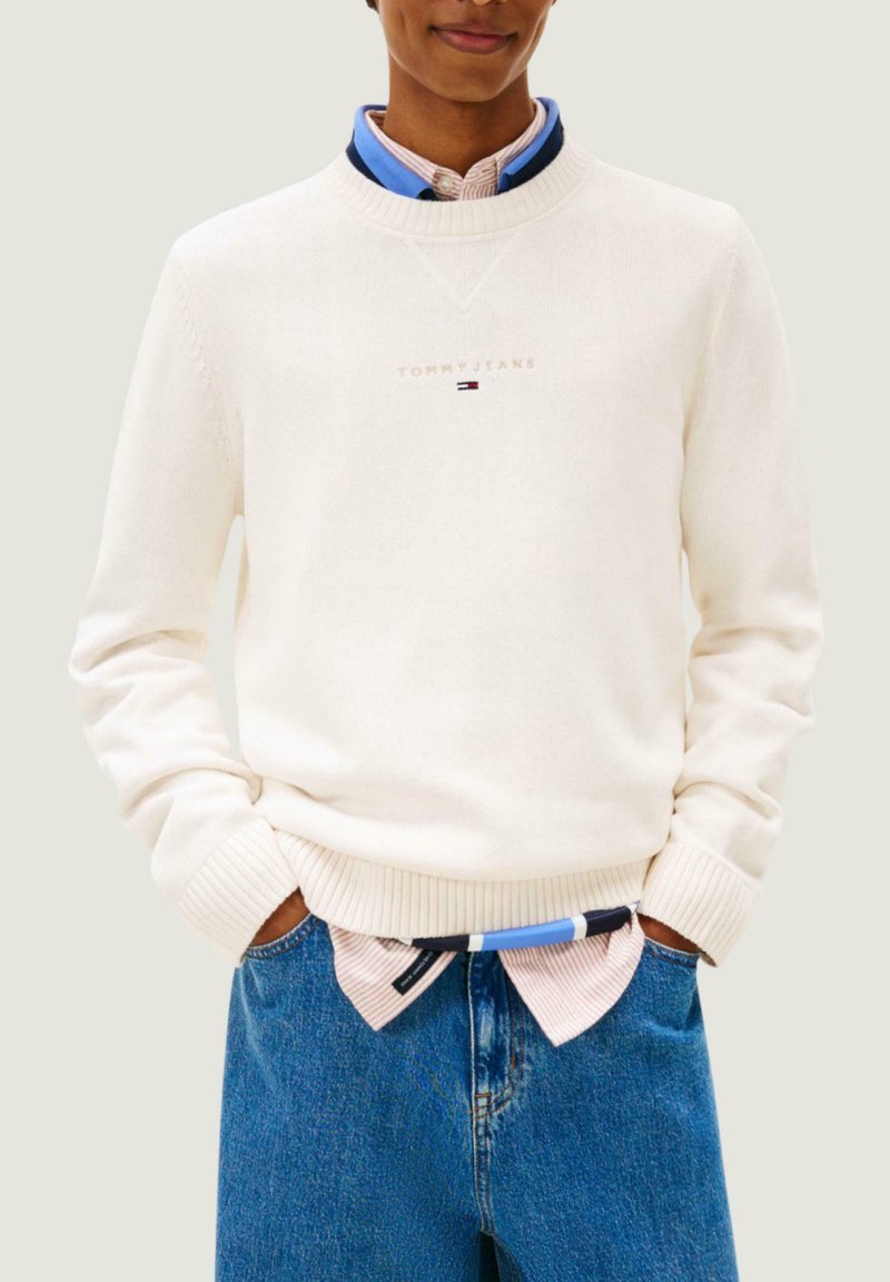 Pull blanc en maille avec poignets et ourlet côtelés, arborant un logo discret sur la poitrine. Associé à une chemise à col rayée bleu et blanc en dessous.