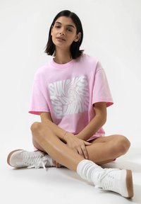 T-shirt surdimensionné rose clair avec un graphique en spirale blanche, associé à des baskets blanches et des chaussettes de cheville blanches, sur un fond clair.