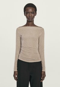 Donna che indossa una maglietta beige a spalle scoperte con maniche lunghe e pantaloni neri a gamba larga, in piedi contro uno sfondo chiaro e semplice.