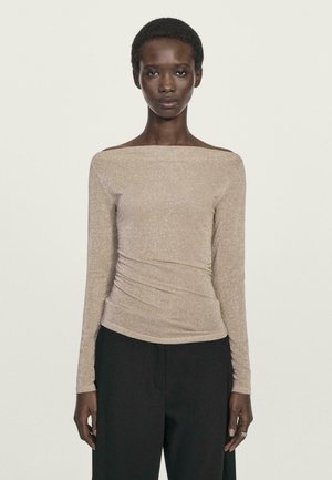 Donna che indossa una maglietta beige a spalle scoperte con maniche lunghe e pantaloni neri a gamba larga, in piedi contro uno sfondo chiaro e semplice.