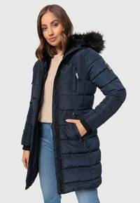 Donkerblauwe gewatteerde jas met capuchon, voorzien van een zwarte nepbont rand, ritszakken, geribde manchetten en een doorgestikt design. Draag met een beige trui en jeans.