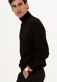 Pull noir à col roulé avec un design ajusté, un col côtelé et une texture lisse. Associé à une montre au cadran blanc et au bracelet noir.