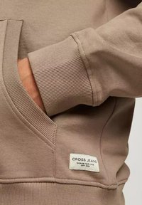 Jasnobrązowa bluza z ściągaczami i kieszenią kangurką, z metką "CROSS JEANS". Materiał wydaje się miękki i teksturowany.