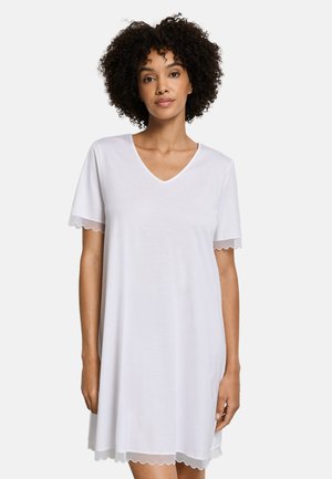 Vrouw met krullend haar draagt een witte V-hals jurk met korte mouwen, afgewerkt met geschulpte kanten randen aan de mouwen en zoom, tegen een effen achtergrond.