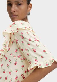 Bloes met bloemenprint, korte gerimpelde mouwen, crème achtergrond en rode bloemen; lichte stof met een zachte textuur en delicate frills.