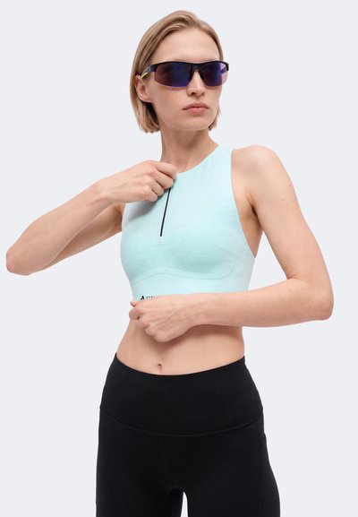 Frau trägt blaues ärmelloses Sporttop und schwarze Leggings, stellt den vorderen Reißverschluss ein und trägt dunkle Sonnenbrille vor hellem Hintergrund.