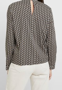 Top lungo nero a maniche lunghe con un motivo geometrico in crema e marrone chiaro, dotato di dettaglio a chiave sulla schiena e texture liscia.