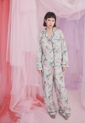 Kobieta stojąca w kwiecistym, jasnoniebieskim komplecie piżamy z długimi rękawami i spodniami, na tle różowo-fioletowej przezroczystej tkaniny.