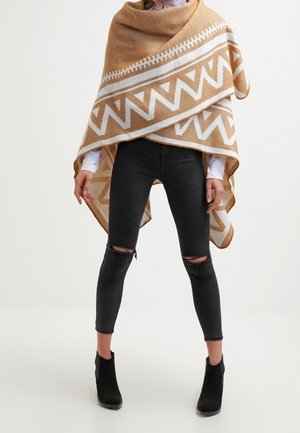 Henkilö, joka pukeutunut beige-valkoiseen geometriseen kuvioituun ponchoon, mustiin revittyihin skinny-farkkuihin, valkoiseen pitkähihnaiseen paitaan ja mustiin nilkkureihin.