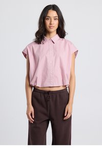 Camicia corta con maniche corte, bottoni frontali, in rosa chiaro con righe verticali bianche. Tessuto morbido, design con colletto, abbinata a pantaloni della tuta marrone scuro.