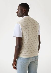 Beige gestrickte Weste mit einemDiamantmuster und Logoakzenten, geripptem Saum und Armöffnungen. Getragen über einem weißen T-Shirt, kombiniert mit hellen Jeans.