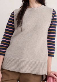 Beige Wolle ärmelloser Pullover mit geripptem Saum, getragen über einem gestreiften Langarmshirt mit navy, pink und senfgelben Streifen.