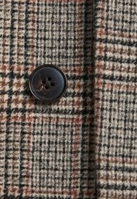 Tessuto di lana con un motivo a quadri beige, marrone e nero, con un bottone marrone scuro a quattro fori ben in vista.