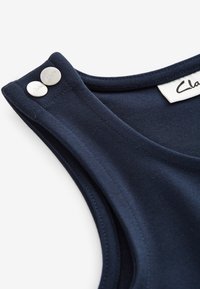 Tengerészkék ujjatlan ruhadarab közeli kép, amelyen a vállpántok láthatók, rajtuk két ezüstszínű patent, amelyeken a "Clarks" felirat található, valamint a nyakkivágás címkéje.