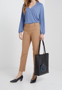 Blouse bleu à manches longues, pantalon beige ajusté, sac fourre-tout noir avec un motif de lapin, et escarpins en vernis bleu marine à talons carrés.