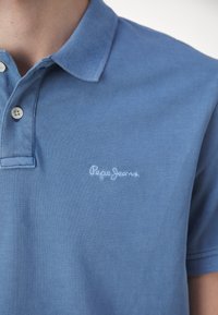 Gros plan d'un polo bleu avec deux boutons blancs et un logo "Pepe Jeans" légèrement brodé sur la poitrine gauche.