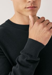 Suéter negro de punto con cuello y puños acanalados, con tejido texturizado. Primer plano de una mano apoyada en la barbilla sobre un fondo neutral.