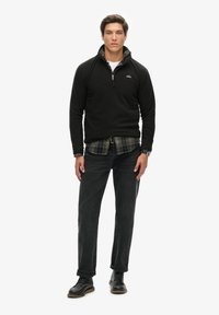 Pullover in pile nero con colletto alto, camicia a quadri sotto, jeans scuri e scarpe nere stringate, in piedi su uno sfondo bianco.