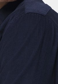 Námornicky modrá kordurová košile s texturovaným povrchem, která má špičatý límec a viditelný žebrovaný vzor na látce.