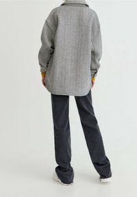 Personne debout de dos portant une veste en arêtes de poisson grise, un pantalon ample foncé aux ourlets effilochés et des baskets blanches sur un fond blanc.