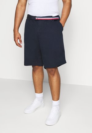 Dunkelblaue Baumwollshorts mit Seitentaschen, einem gewebten Gürtel mit rot-weißen und blauen Streifen, kombiniert mit weißen Sneakers und Kniestrümpfen.