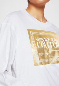 Bílá dlouhá tričko s dlouhým rukávem, které má zlatě tištěné logo a dekorativní lem. Materiály vypadají měkce a mají hladkou texturu.